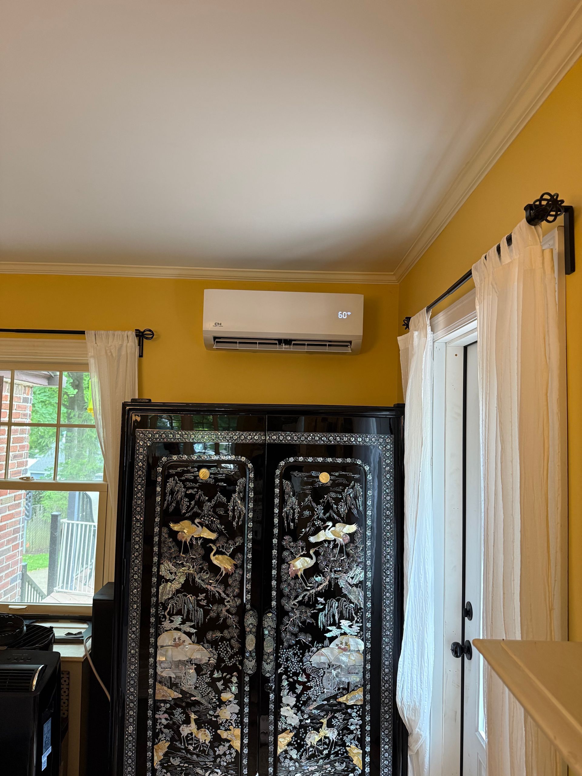 Yellow room with air conditioner above a decorated black wardrobe and white curtains by a window