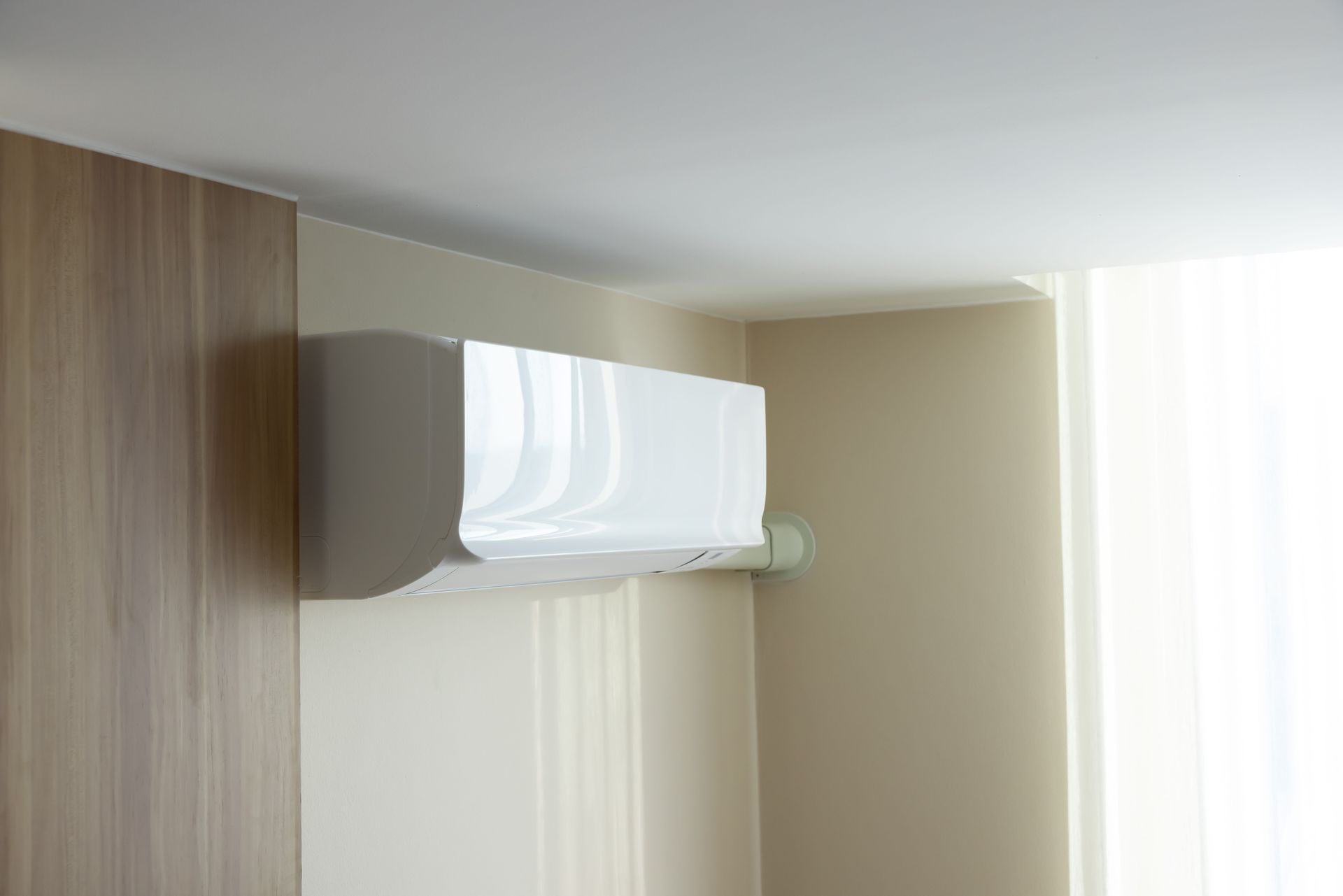 Wall-mounted white air conditioner near a window with sheer curtains in a beige room