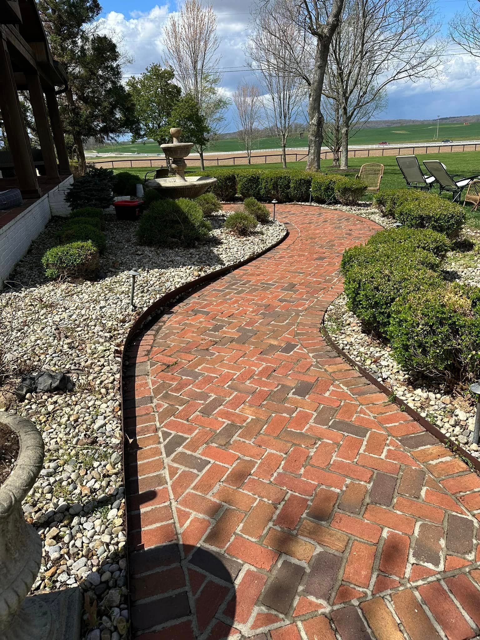 Brick pathway curving through a landscaped garden with trimmed hedges, a fountain, and gravel border.