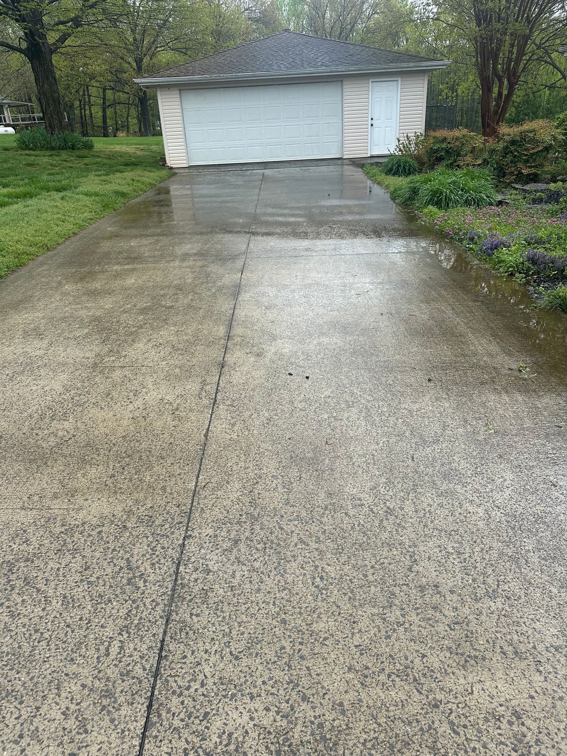 Wet concrete driveway leading to a garage with closed white doors. Green grass and trees frame the scene.