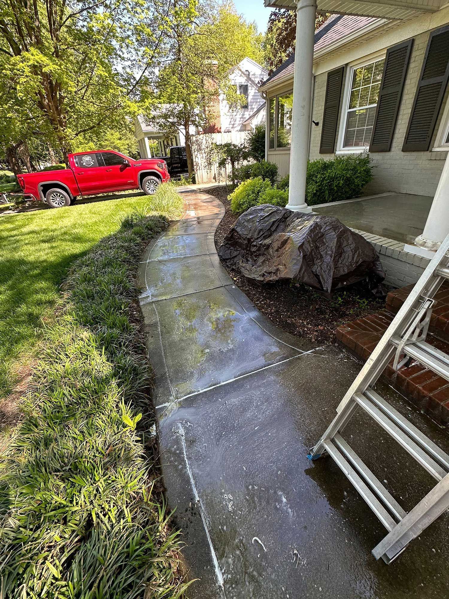 A brick-lined, concrete walkway being power-washed; a large rock sits near the porch of a home, and a red truck is parked in the yard.