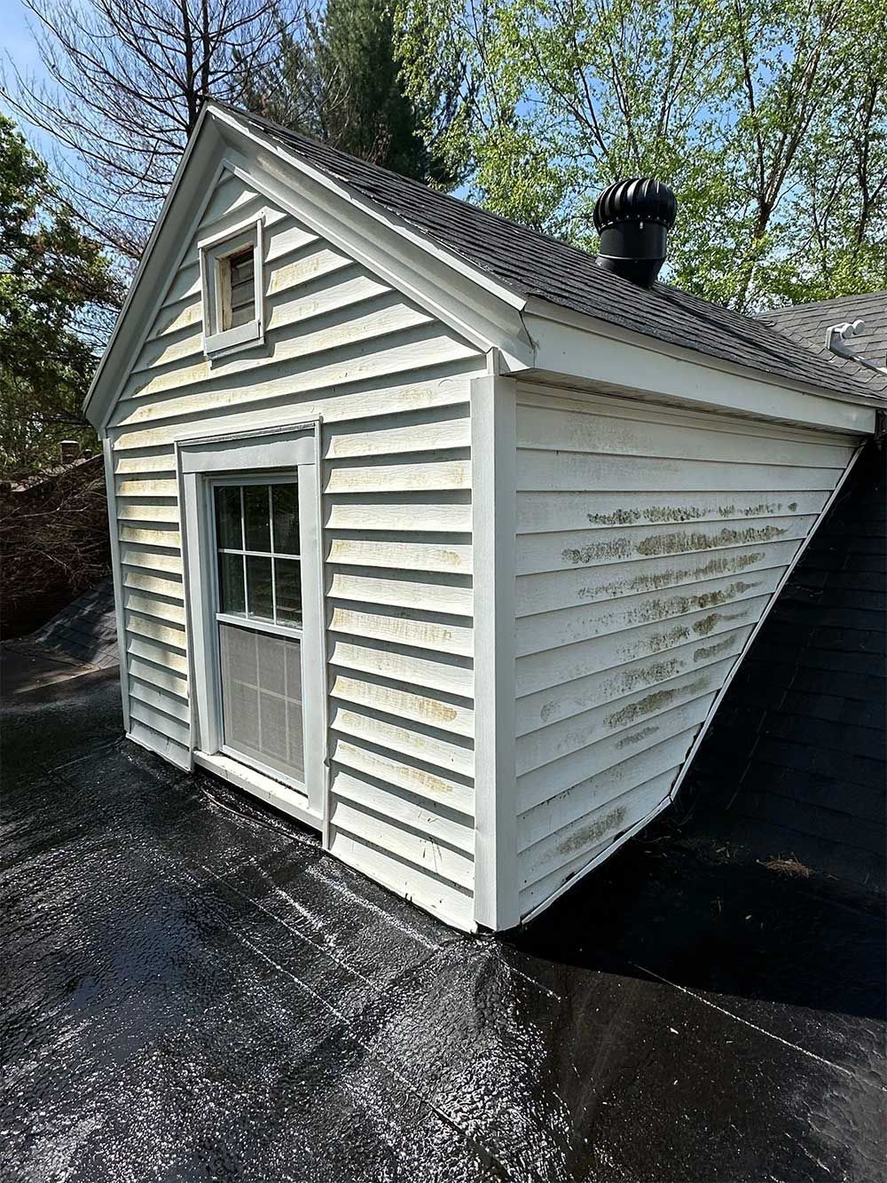 Small, white shed-like structure on black roofing. Shows siding, door, window, and a chimney on a sunny day.