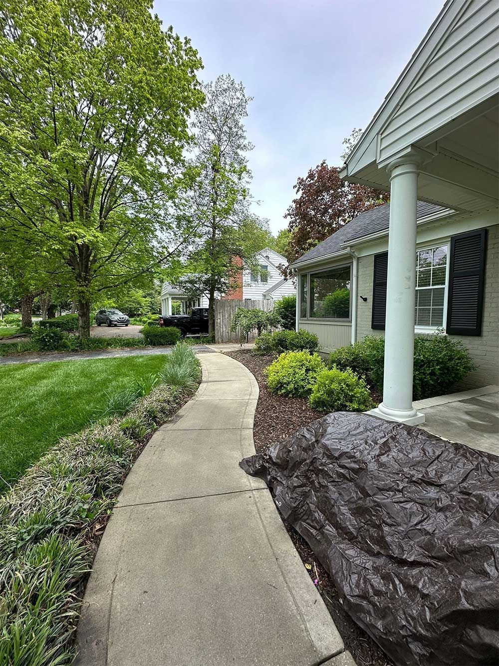 Sidewalk leading to a house with a porch and landscaping. Green grass and trees surround the walkway.