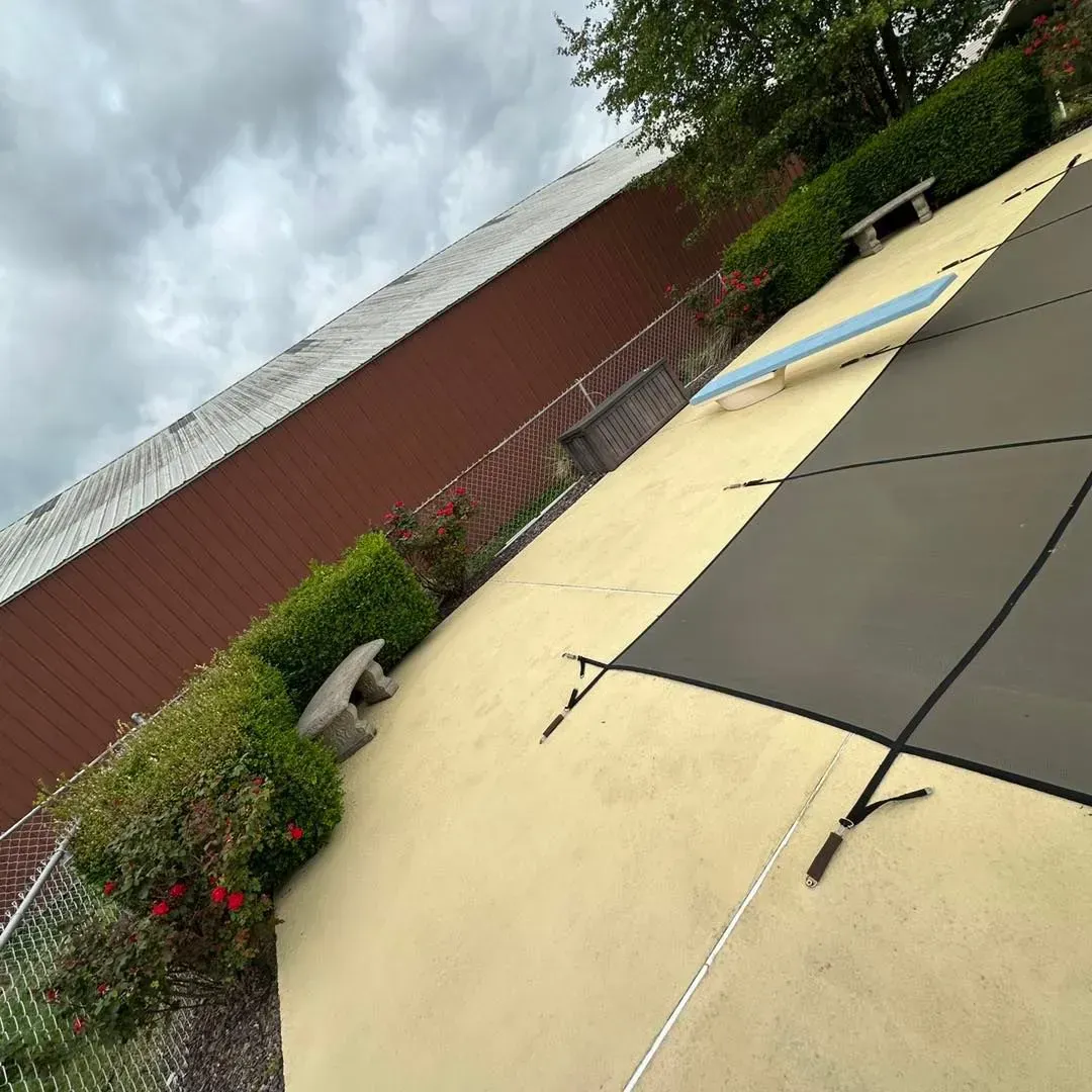 Pool covered with black and beige tarp. Brown building and green hedges in the background. Cloudy sky.