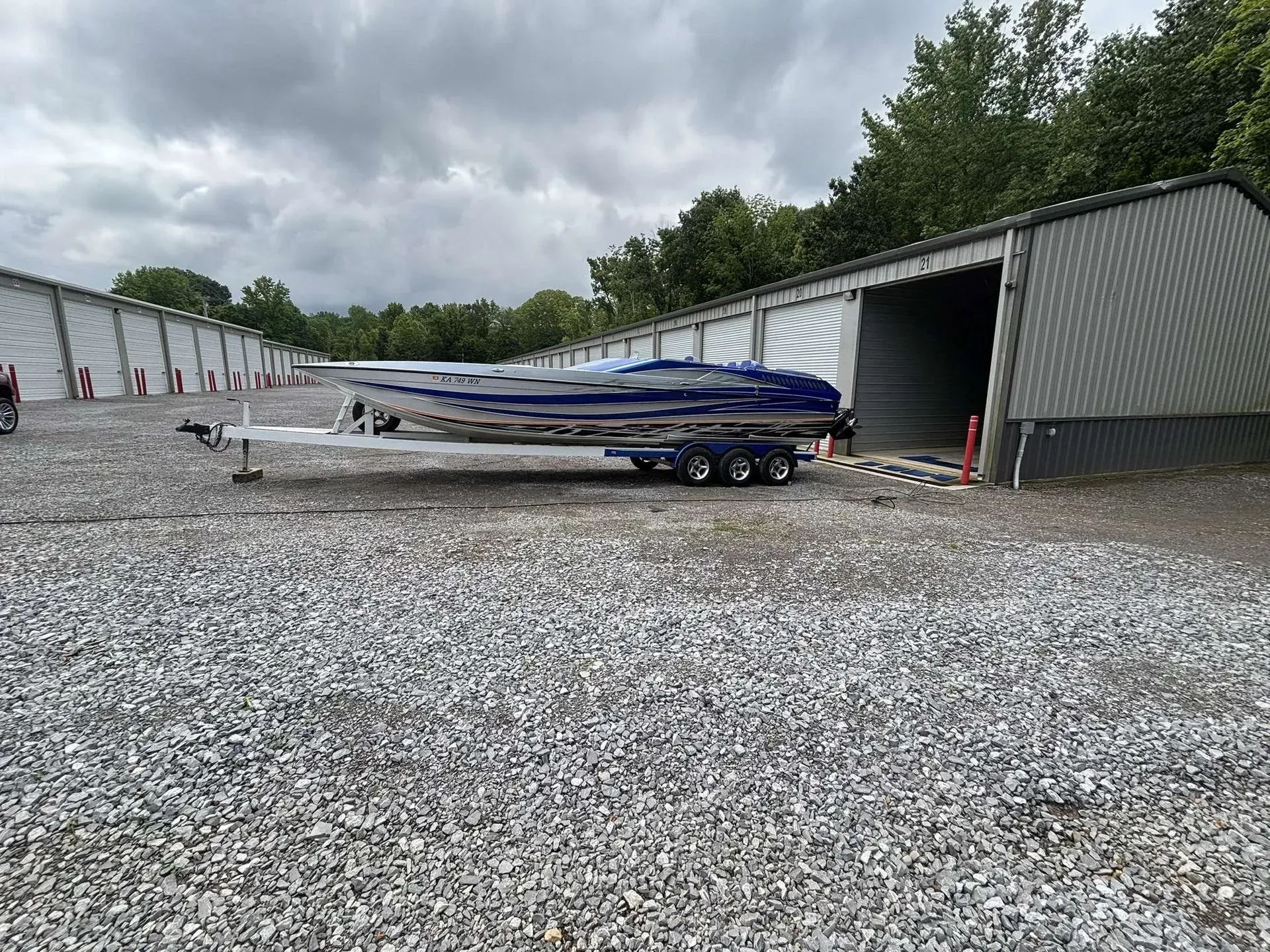 A blue and silver speedboat on a trailer outside a storage unit building on gravel.
