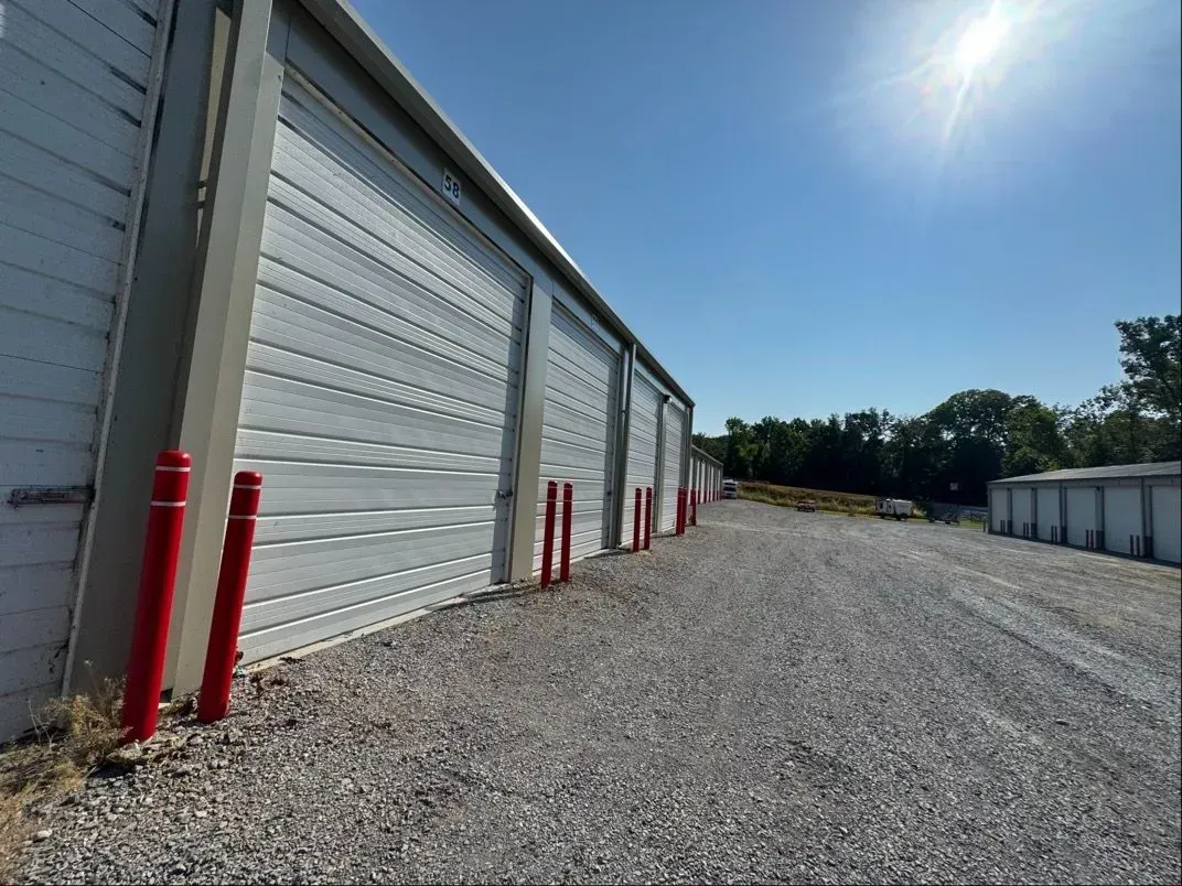 Row of white storage unit doors with red bollards along a gravel driveway under a bright blue sky.