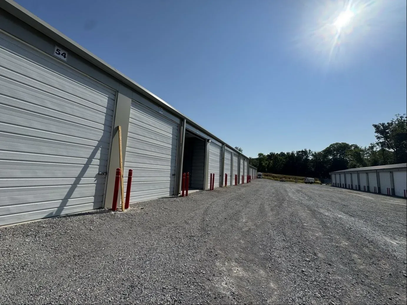 Storage units with open and closed doors under a bright sun. Gravel driveway.