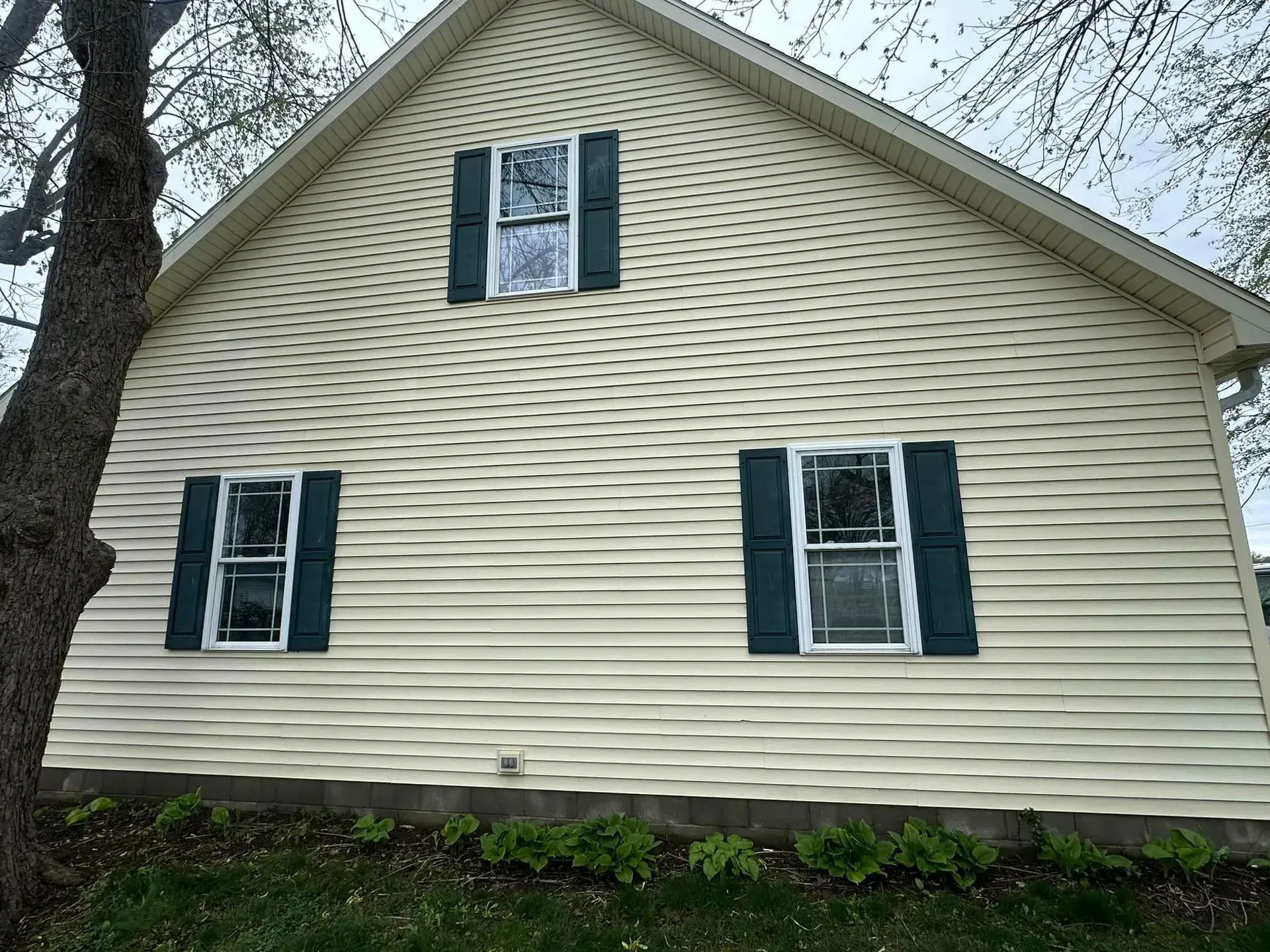 Beige house with green shutters, tree on left, blue sky.