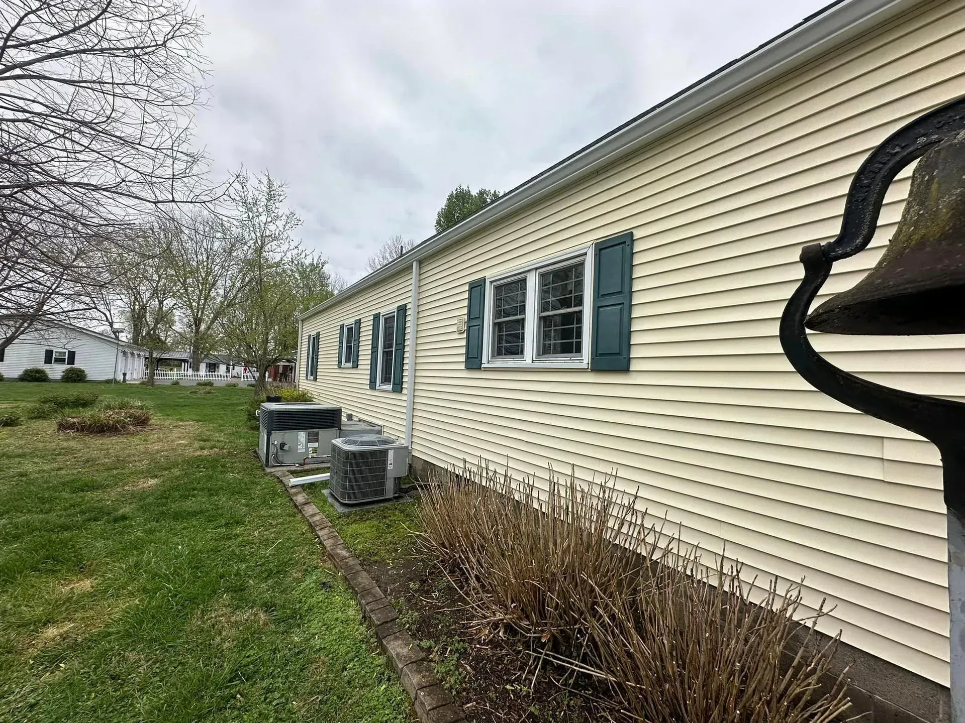 Side of a light yellow building with dark green shutters.  A bell is in the foreground, with grass and trees visible.