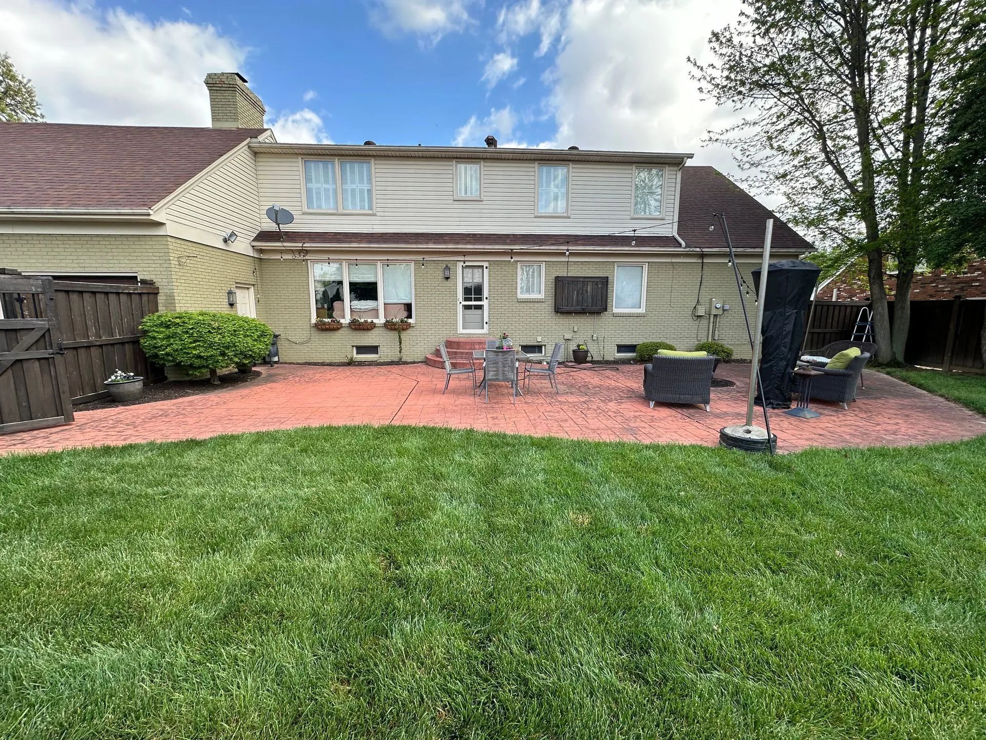 Backyard with red patio, chairs, and house with brown roof. Green grass and trees under a cloudy sky.