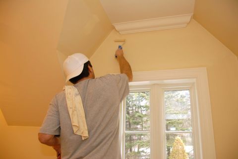 Person painting a cream-colored wall near a window, reaching up with a roller.