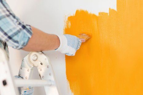 A person wearing a glove and plaid shirt uses a brush to paint a wall with vibrant yellow paint while standing on a ladder.