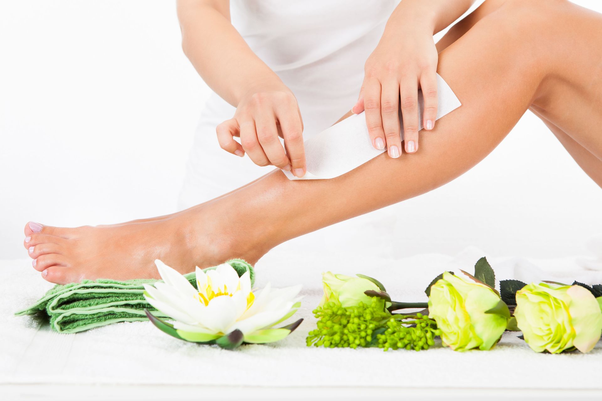 Woman waxing leg with wax strip, flowers, and towels on a white surface.