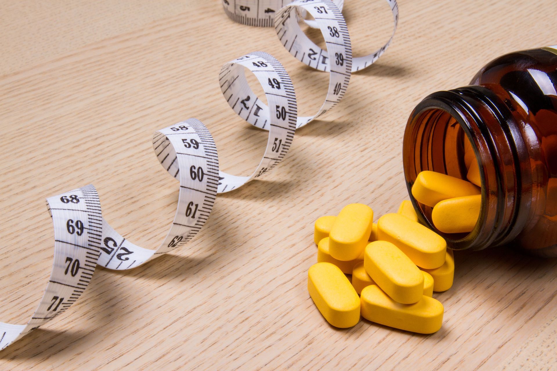 Yellow pills spilling from a brown bottle, with a white tape measure on a wooden surface.
