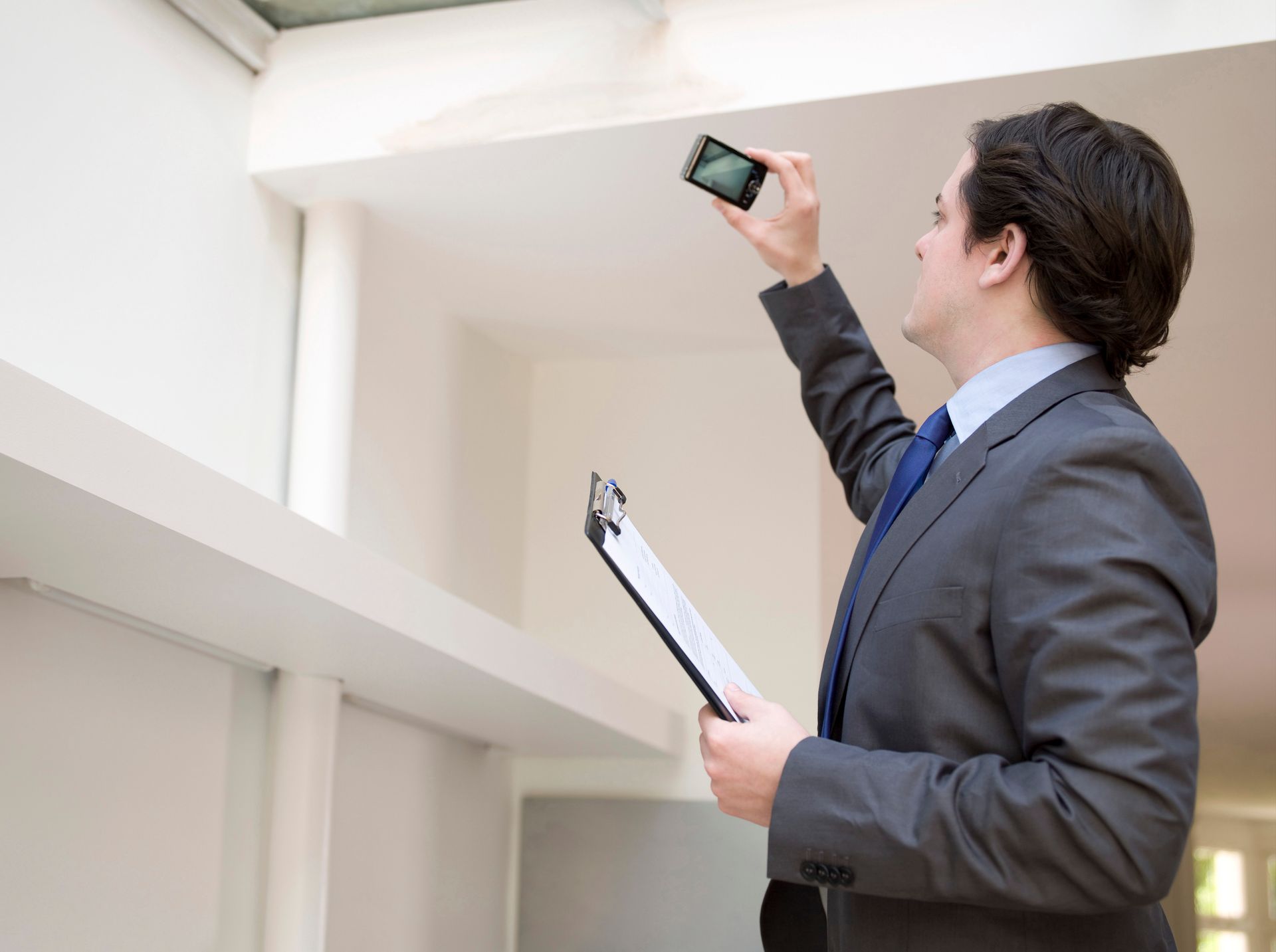 Man in suit inspects ceiling with phone and clipboard.