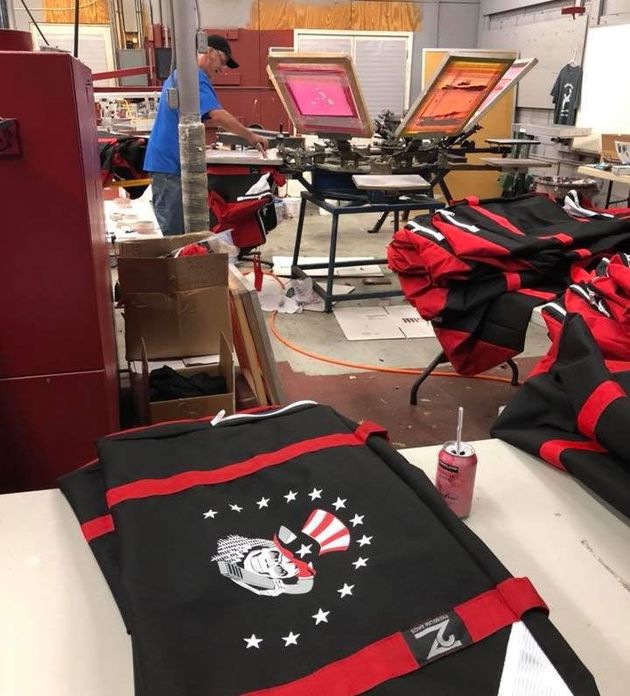 A screen printing workshop featuring a worker at a press and stacks of black and red custom-printed fabric items.