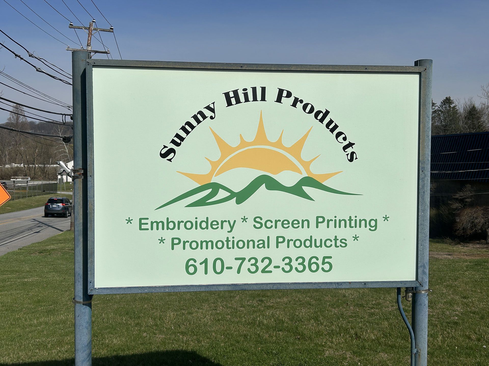 Sign for Sunny Hill Products with a sun and hill logo, offering embroidery, screen printing, and promo products. 610-732-3365.
