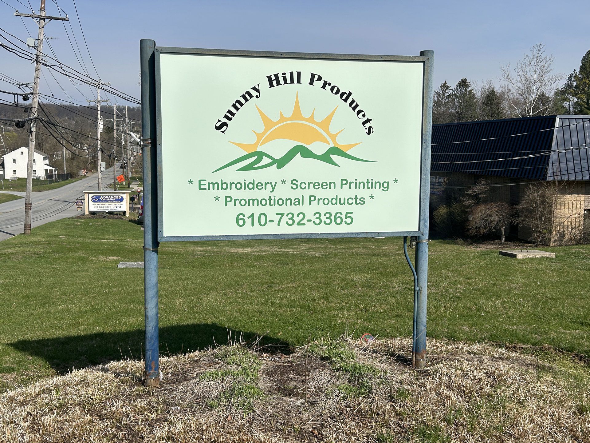 A Sunny Hill Products sign in a grassy area, advertising embroidery, screen printing, and promotional products at 610-732-3365.