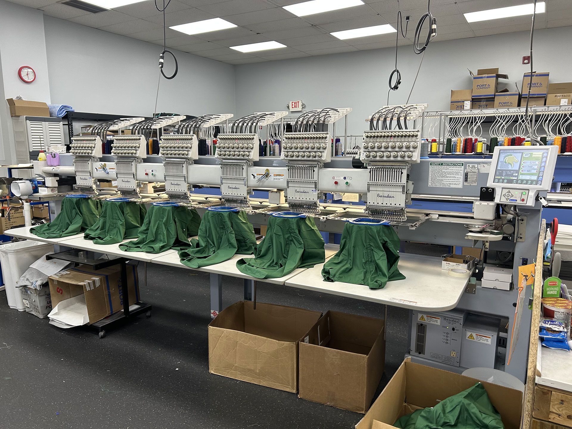 An industrial embroidery machine with multiple heads running on green fabric in a warehouse setting.