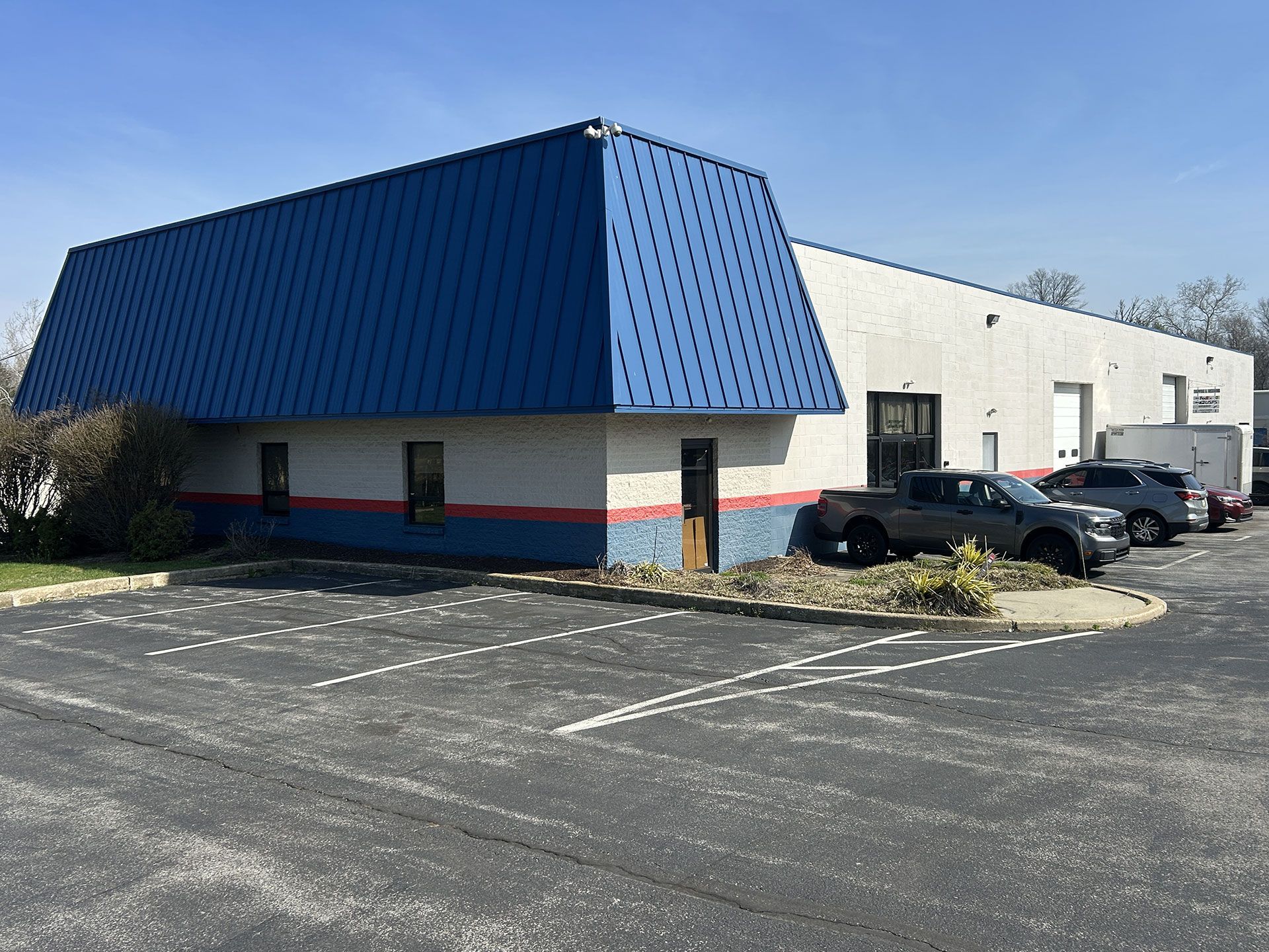 A commercial building with a blue metal mansard roof and a white, single-story exterior, seen from a paved parking lot.