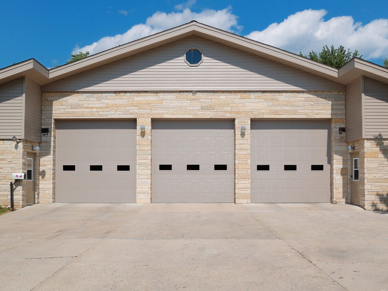 Three beige garage doors on a stone house under a blue sky.