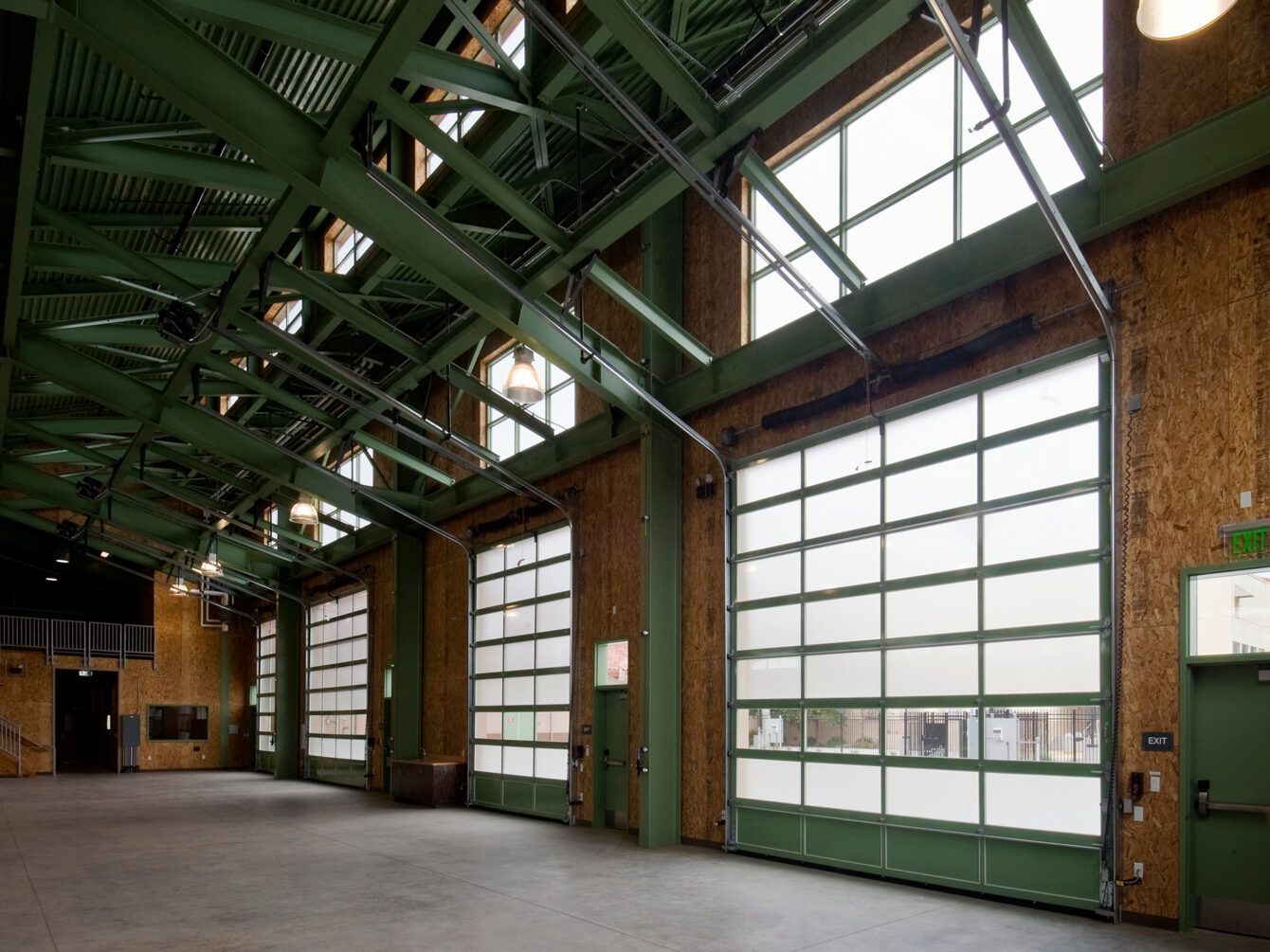 Empty industrial hall with green steel beams, skylights, and large garage doors along one wall
