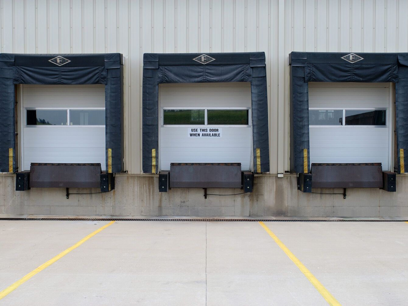 Three loading dock doors with black awnings on a gray warehouse wall and concrete lot