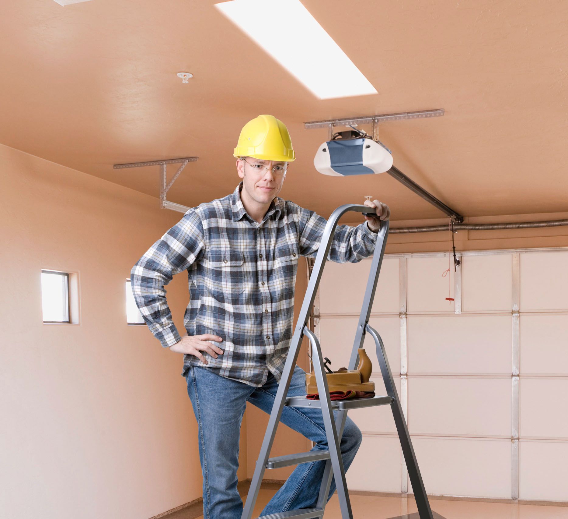 Worker in hard hat standing on a step ladder in a garage, with a garage door opener overhead.