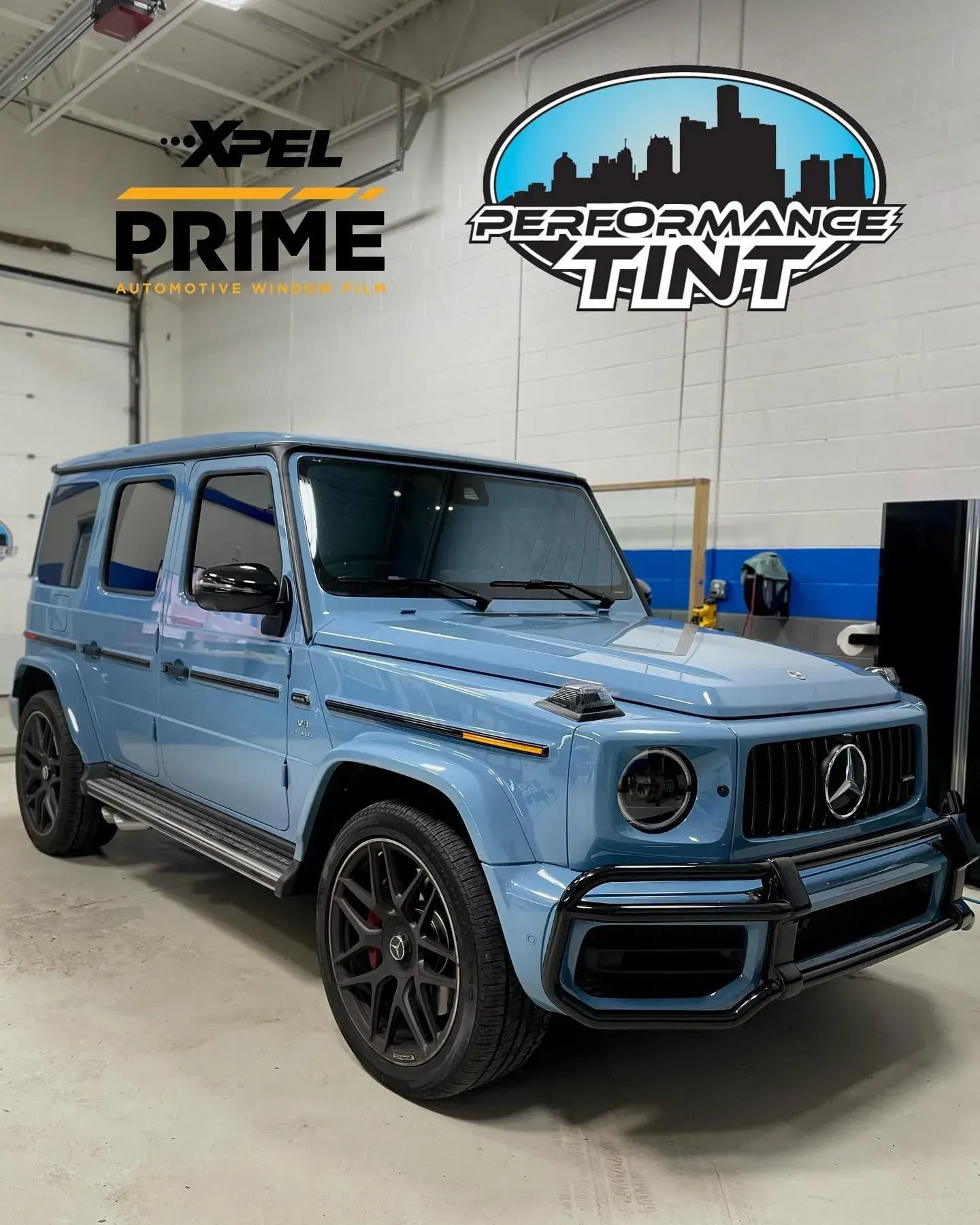 Light blue Mercedes G-Class SUV indoors at a performance tint shop with logo signs on the wall.