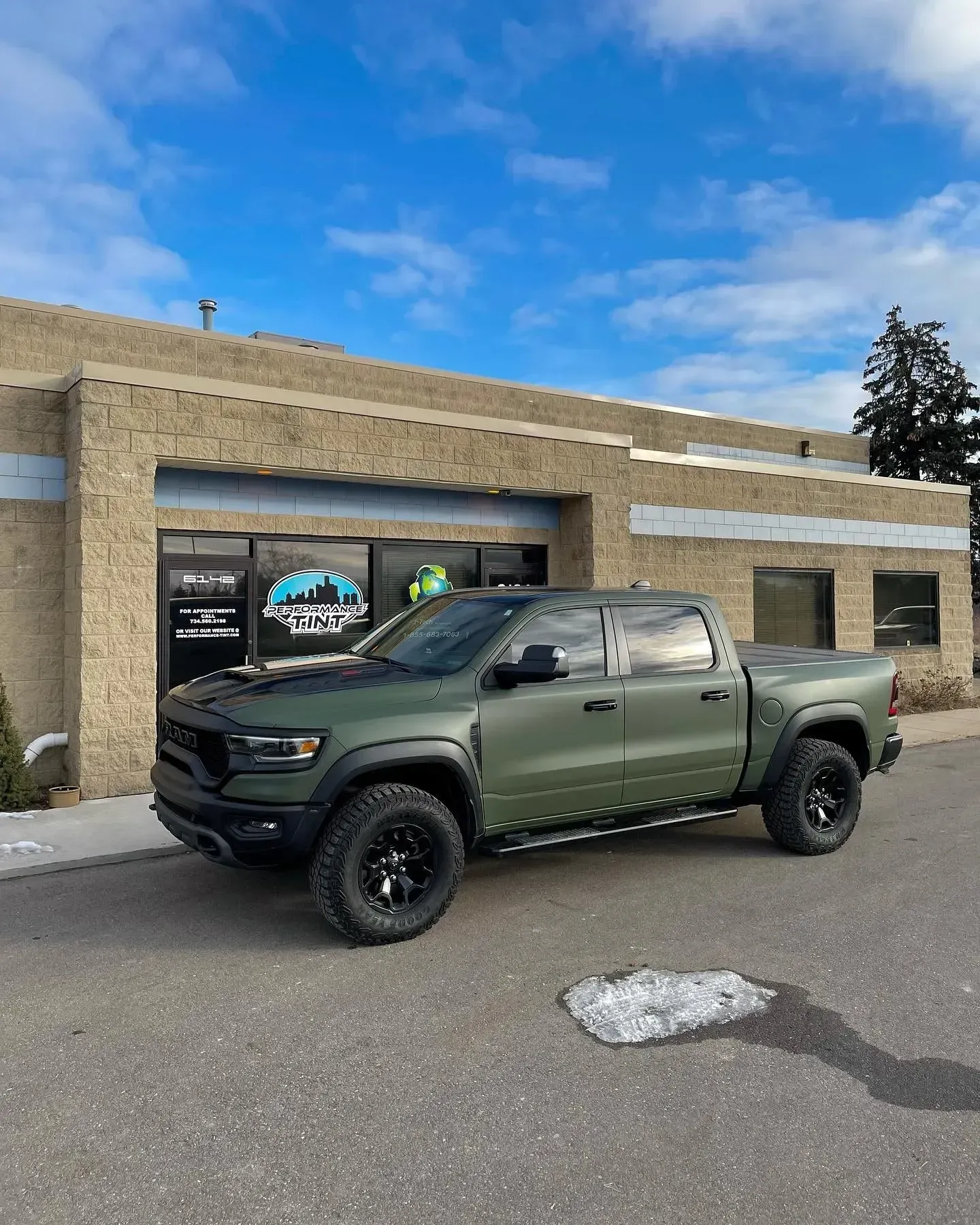 Matte green pickup truck parked outside a tan commercial building under a blue sky.