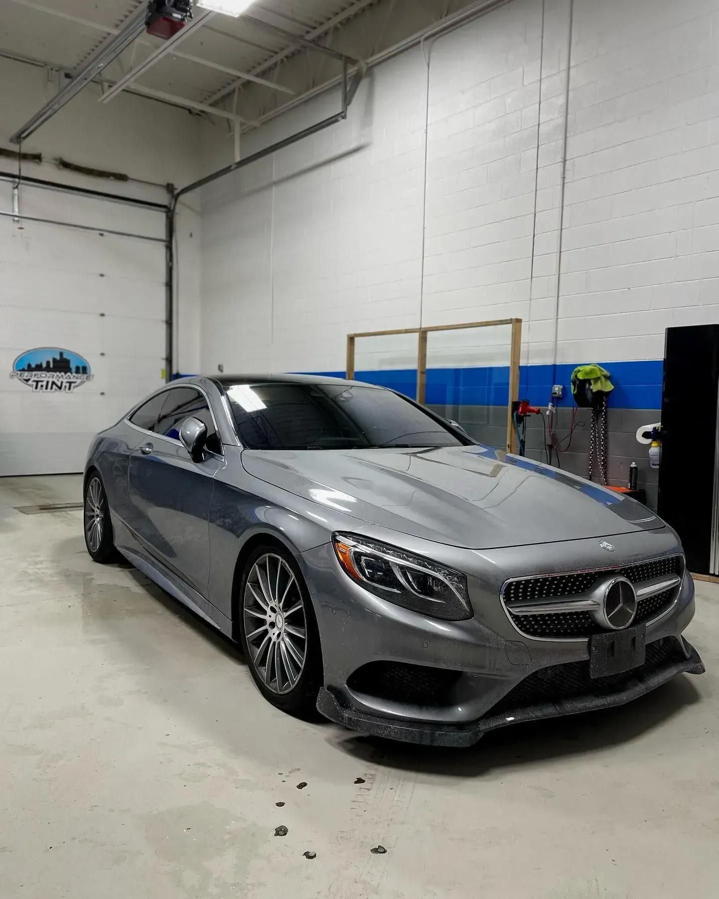 Silver Mercedes coupe in an auto shop, parked indoors near blue walls and a person in high-visibility clothing