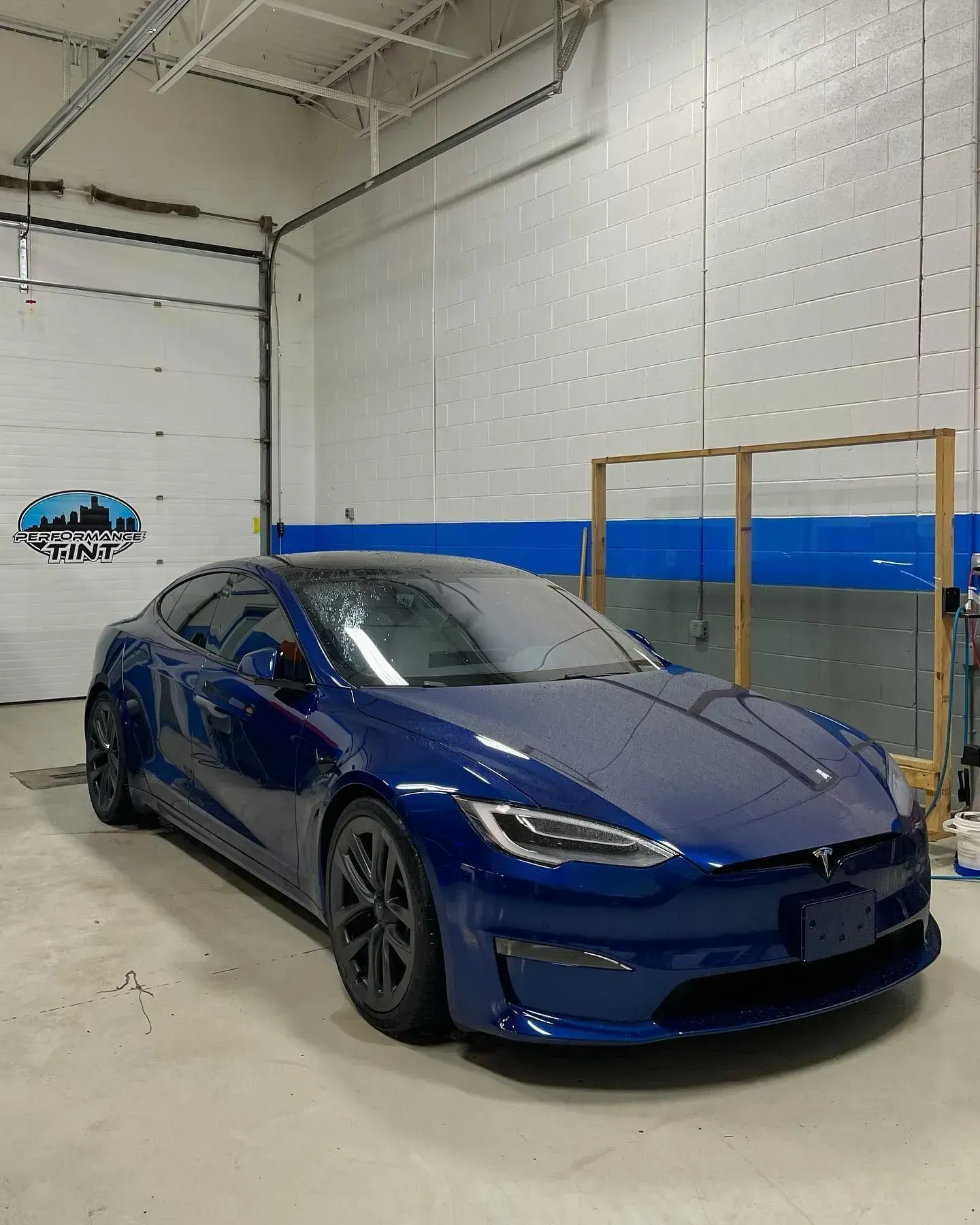 Blue Tesla sedan parked indoors in a garage with white walls and a blue stripe.