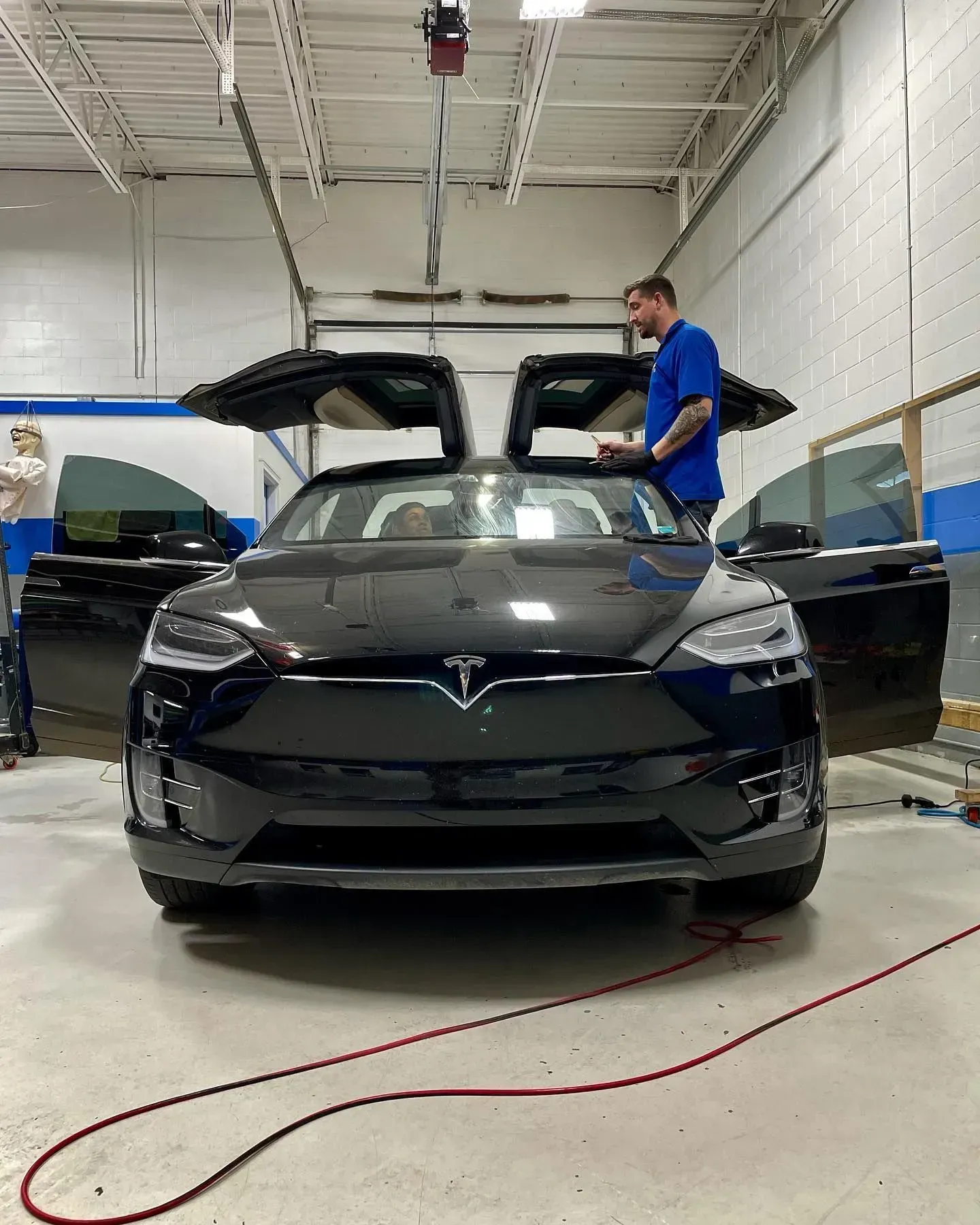 Black Tesla in a workshop with its falcon-wing door raised and a person standing beside it
