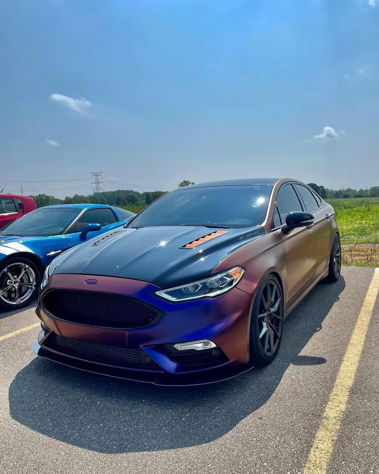 Purple sports sedan parked in a sunny lot, with a blue car beside it and open fields behind.