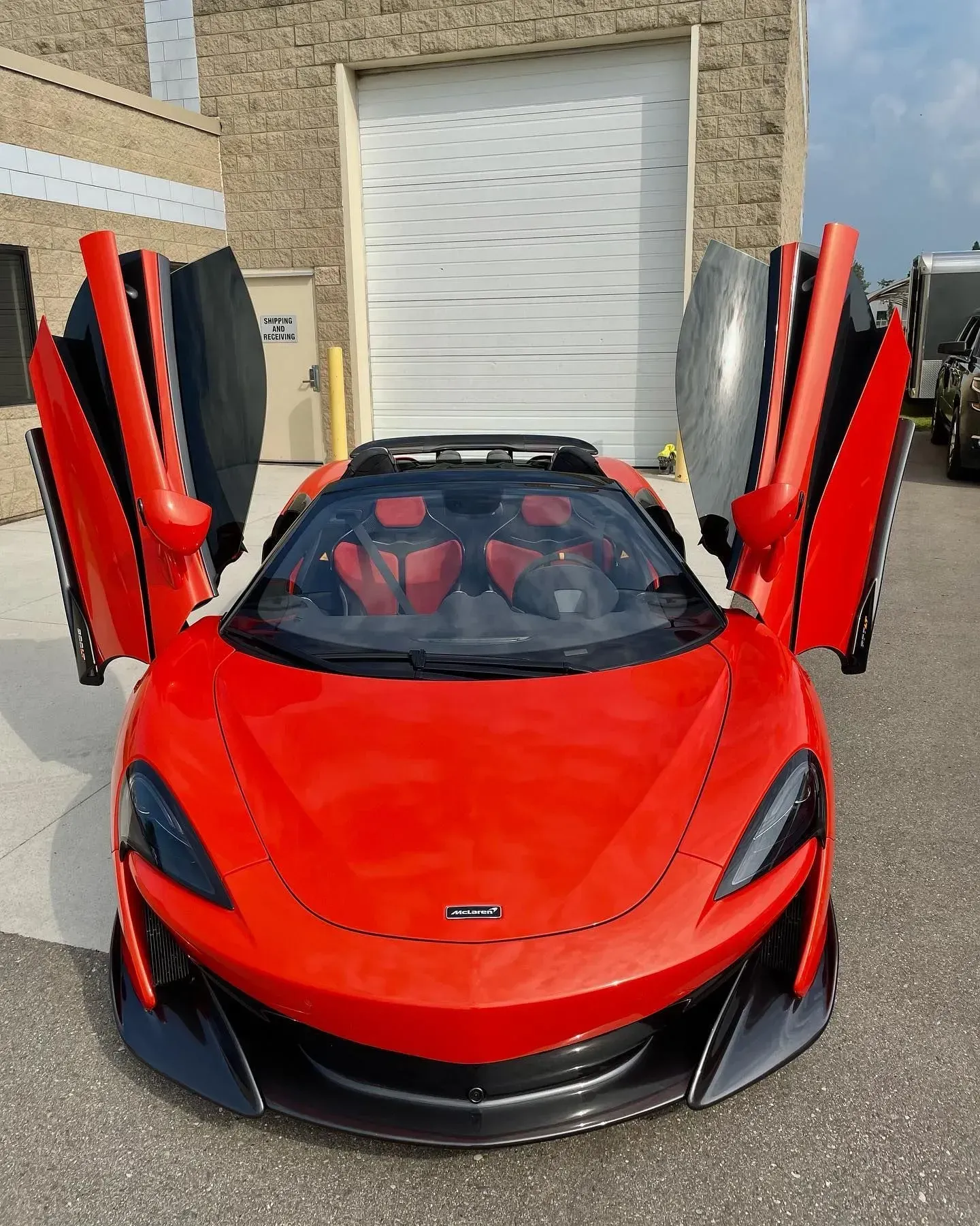 Red McLaren sports car with butterfly doors raised, parked in front of a garage.