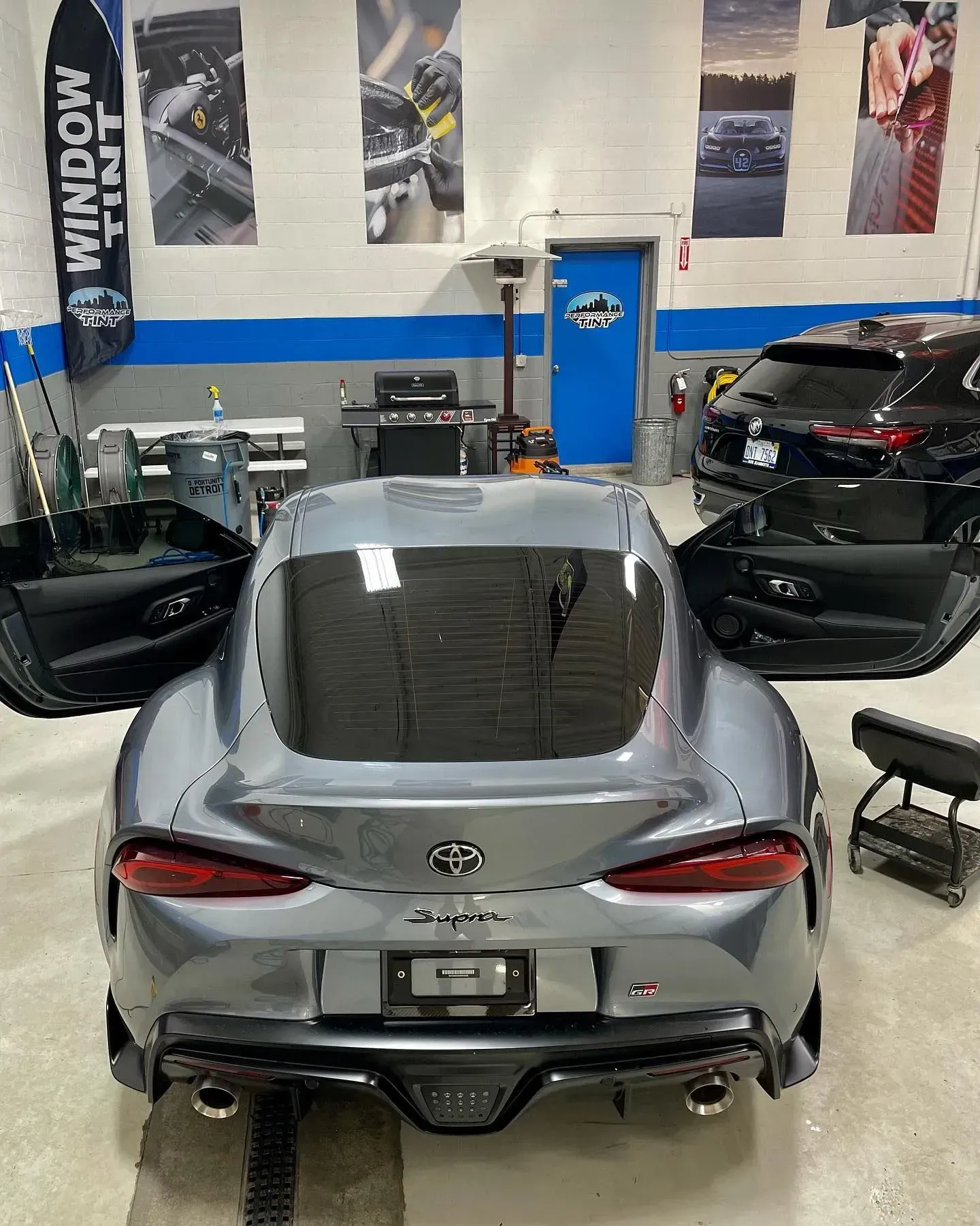Silver sports car in a garage, viewed from the rear with doors open and black cars nearby.