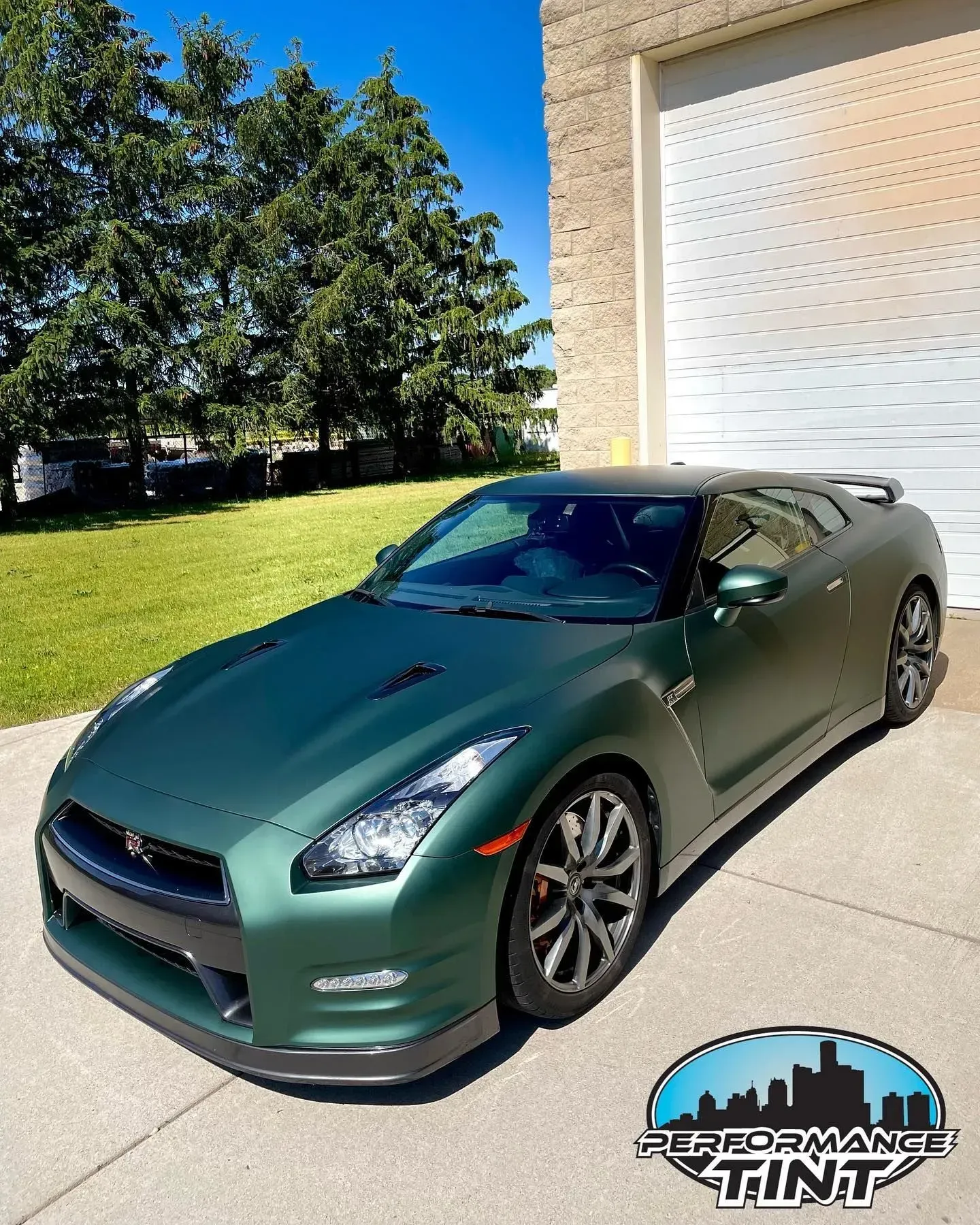 Dark green sports car parked on a driveway beside a building, with trees and lawn in the background.