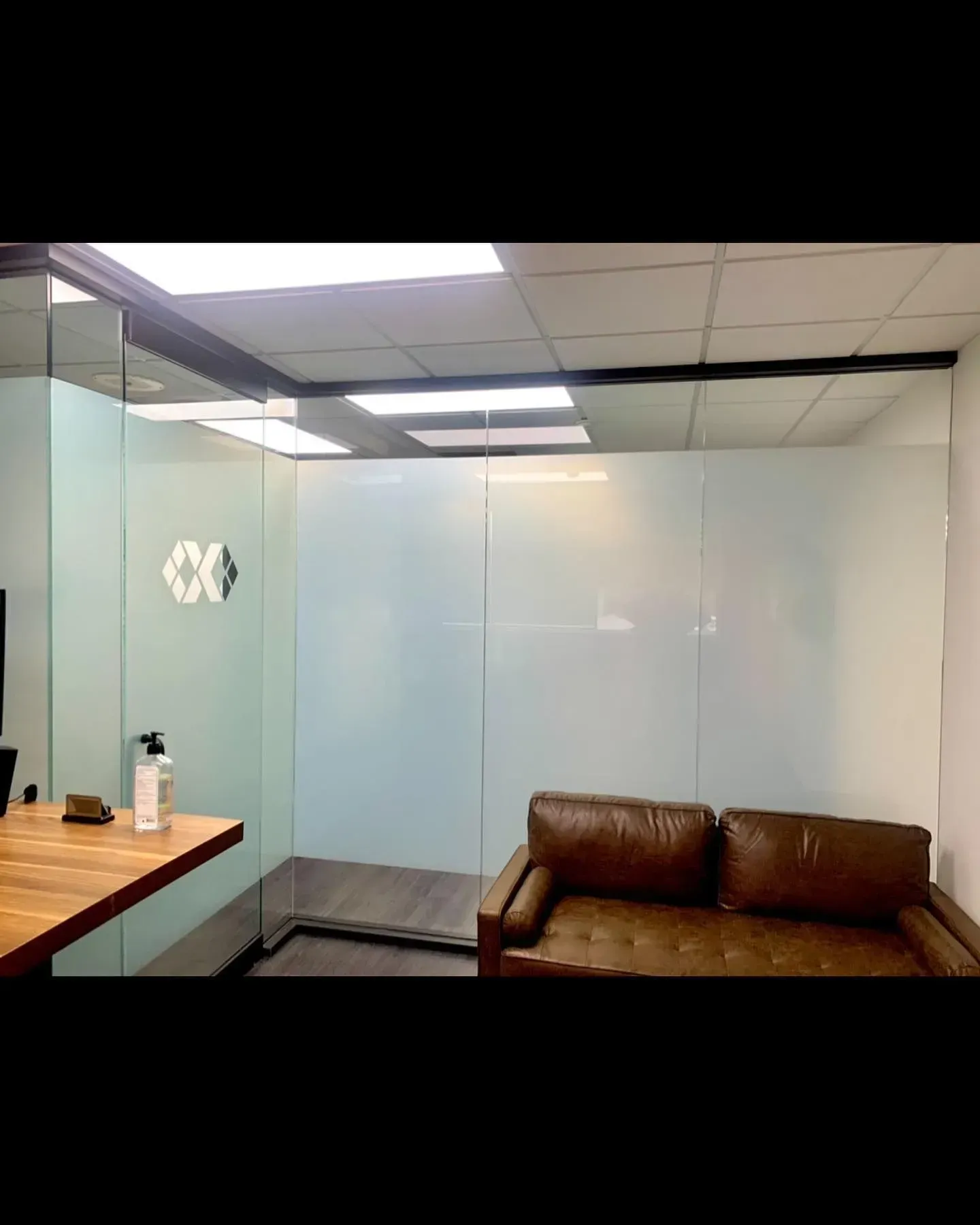 Office waiting area with frosted glass walls, a brown couch, and a wooden table under fluorescent lights