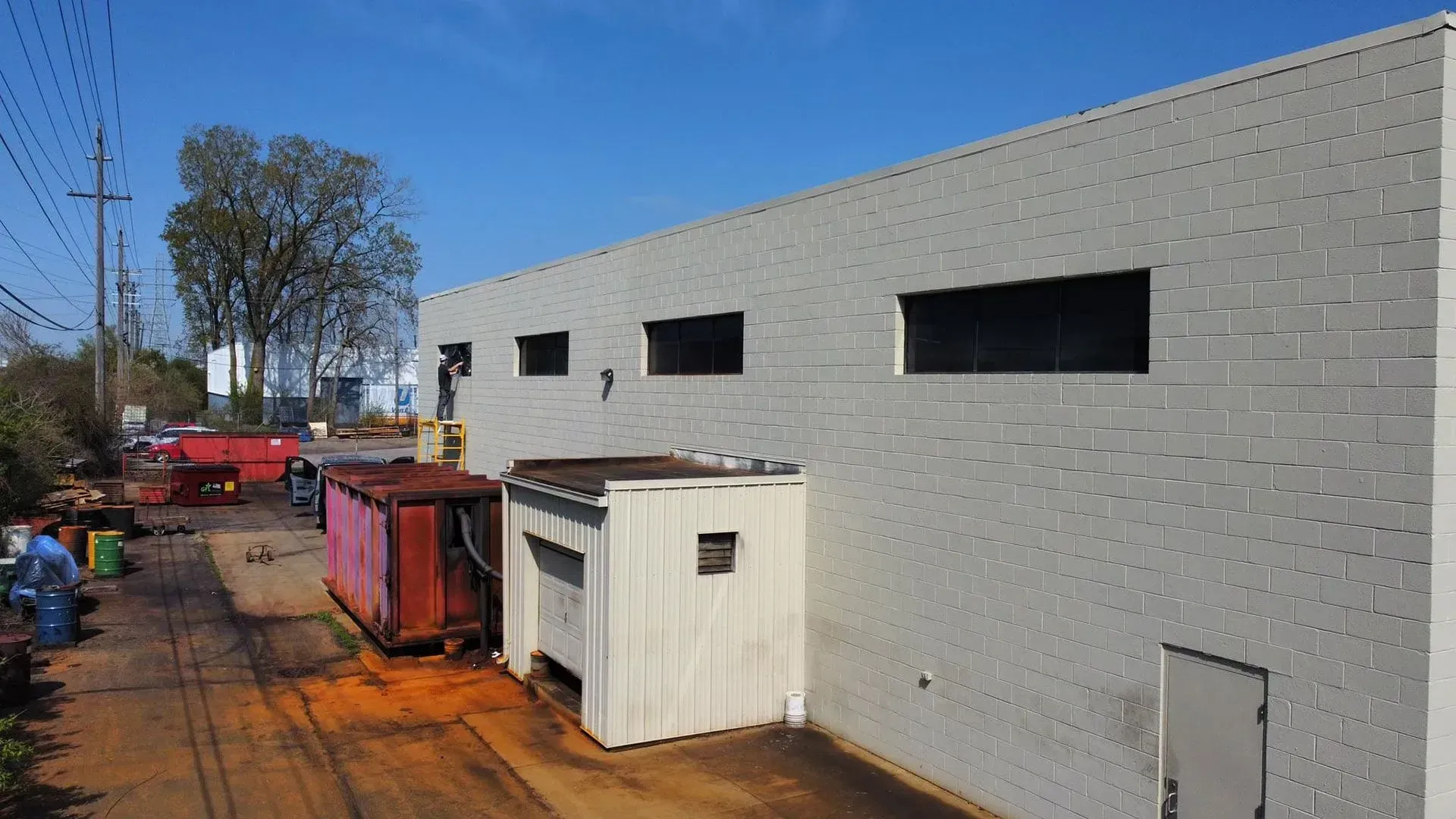 Construction site beside a gray block building with dumpsters, equipment, and a tree-lined path under a blue sky