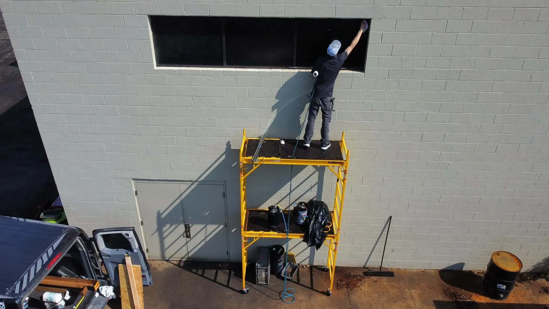 Worker on a yellow lift painting a high window on a gray brick wall.