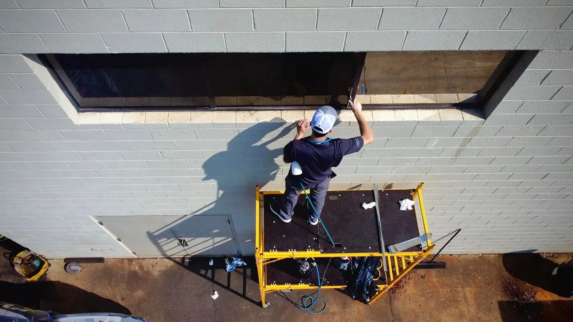 Worker on a yellow platform lifts a window into a gray brick building facade.