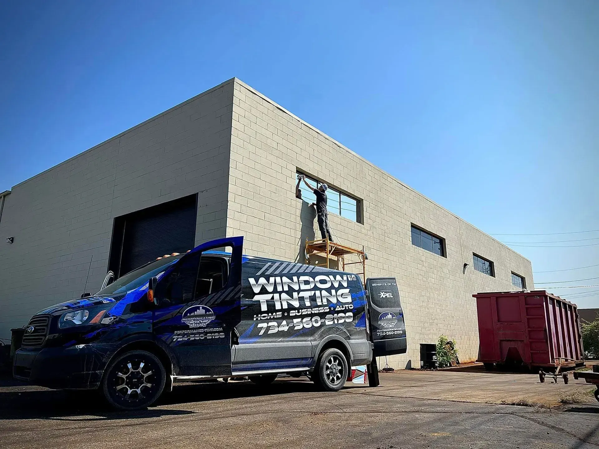 Blue service van parked by a beige warehouse beside a red dumpster under a clear sky