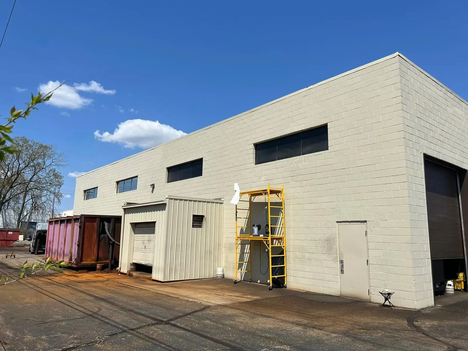 Industrial building with beige walls, small high windows, and containers and ladder outside on a dirt lot