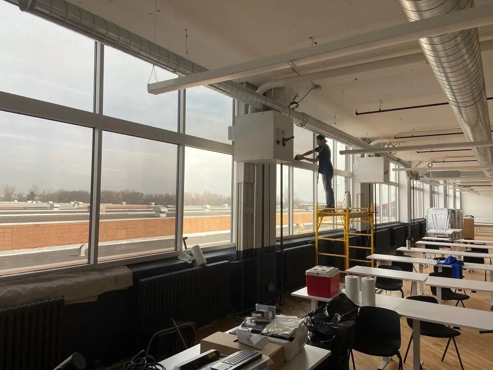 Worker on a ladder installing fixtures in a bright office with large windows and desks.