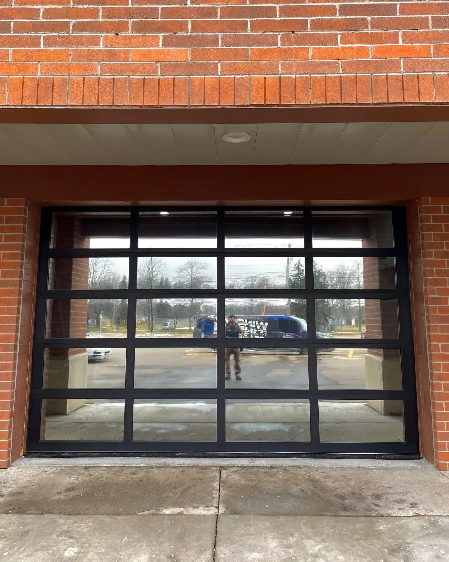 Brick building entrance with black-framed glass garage door reflecting parked cars and street outside