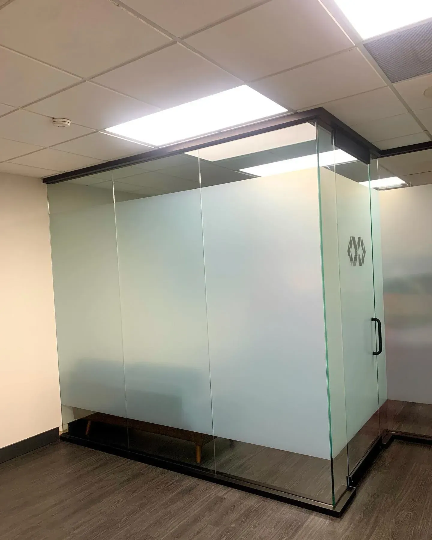 Glass-walled office booth in a hallway with frosted panels and black trim under ceiling lights