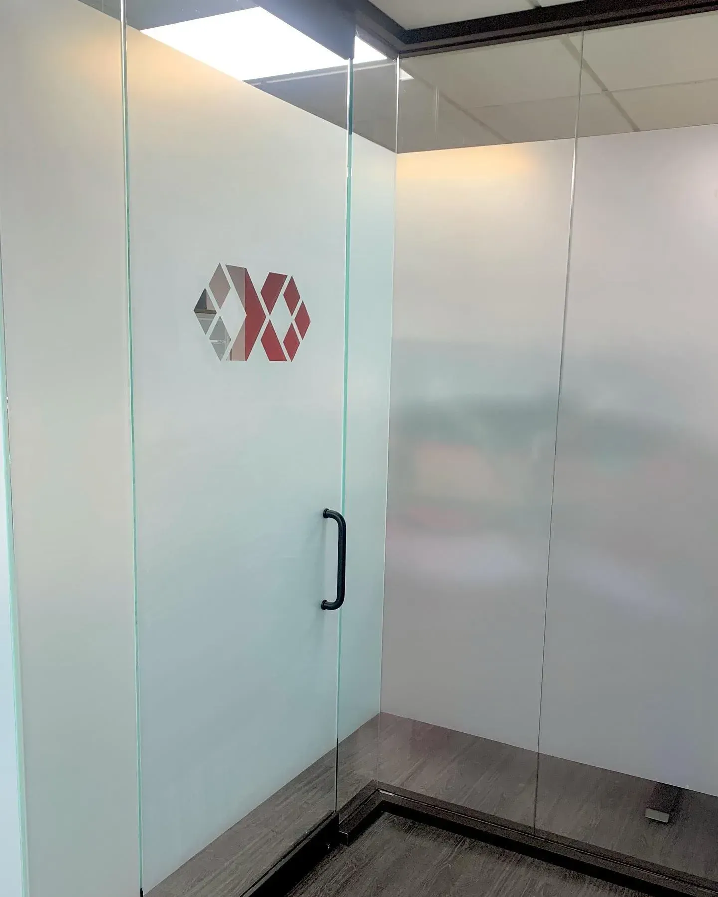 Frosted glass office partition with black handle and geometric logo, in a hallway corner.