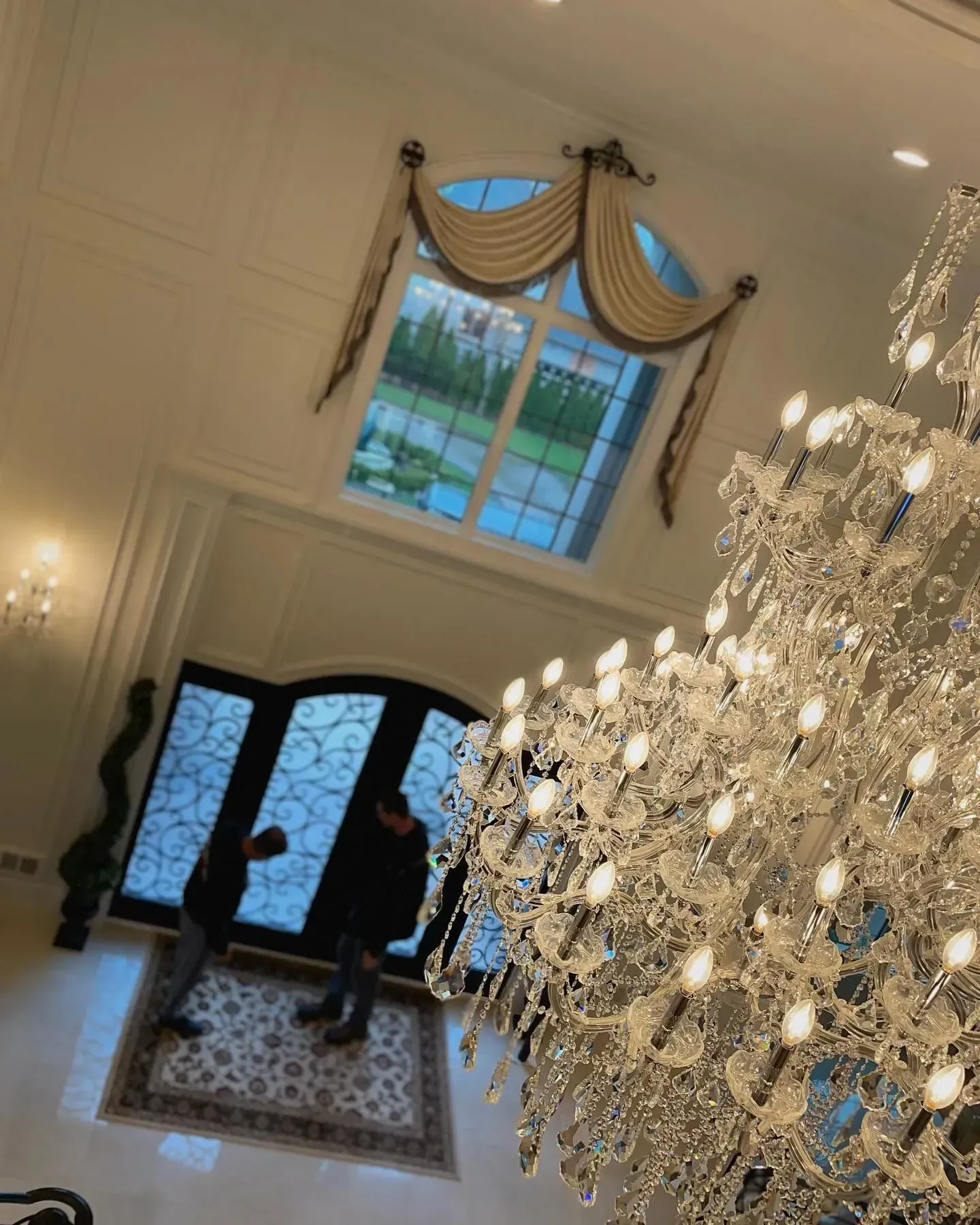 Elegant foyer with a large chandelier, draped window, double doors, and two people near the entrance.