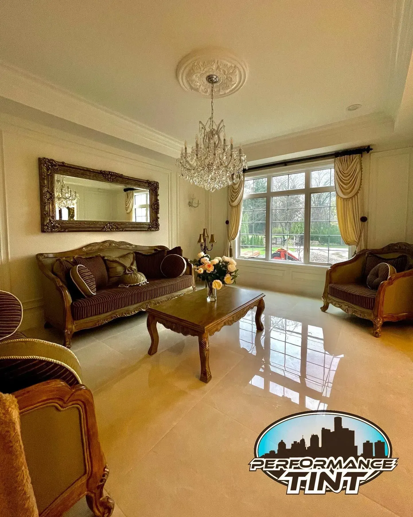 Elegant living room with chandelier, brown sofas, glass coffee table, and large window overlooking greenery