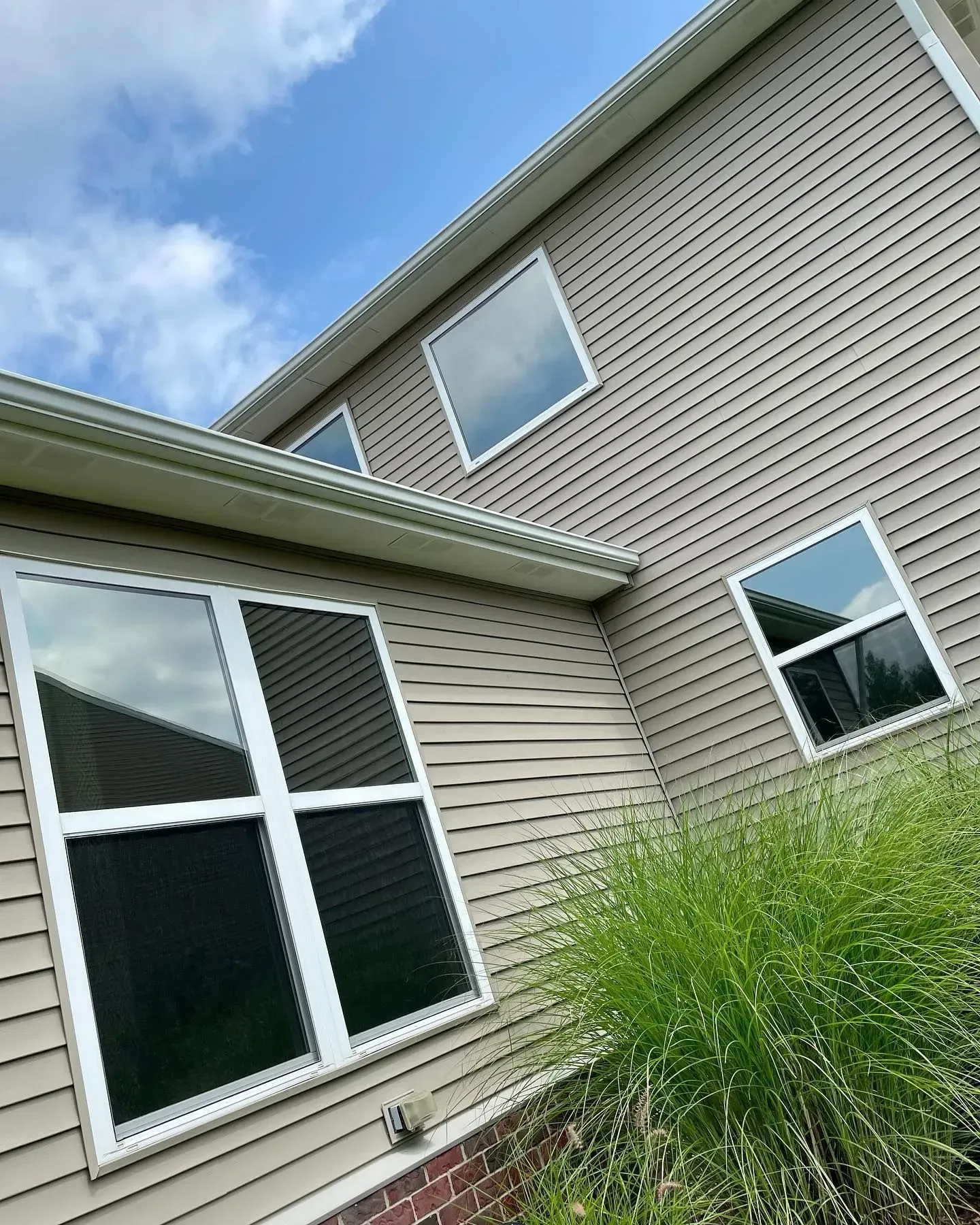 Two-story beige house with white-trim windows and tall green grass in the foreground
